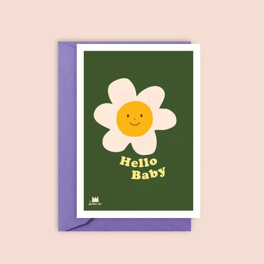 A greeting card with a deep green background featuring a large, illustrated daisy with a smiling yellow centre. The words "Hello Baby" are printed in playful, vintage-style yellow text. The card is paired with a soft purple envelope.