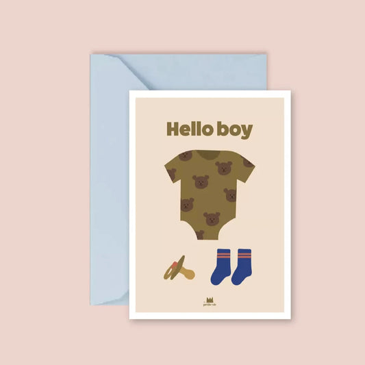 A greeting card with a soft beige background featuring minimalist illustrations of a baby onesie with a bear pattern, a pacifier, and a pair of blue socks. The words "Hello boy" are printed in bold, olive-green text at the top. The card is paired with a pastel blue envelope.