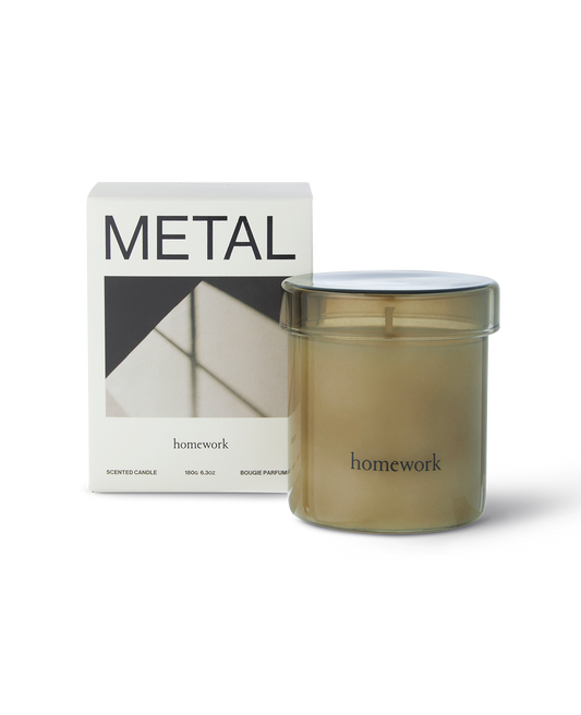 Homework metal scented candle in a moss green glass with matching lid, along the bottom the branding spelling homework in lower case lettering. Next to its packaging, a white box with metal written across the top and a landscape image of shadows on metal below.