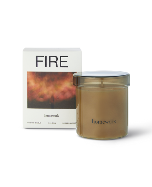 Homework Fire candle in a moss green translucent glass with matching glass lid, with branding of homework along the bottom in lower case text. Next to the packing box, all white with fire spelt across the top, under is a photograph of a blurred fire in red tones. Then the brands name written small underneath the image 