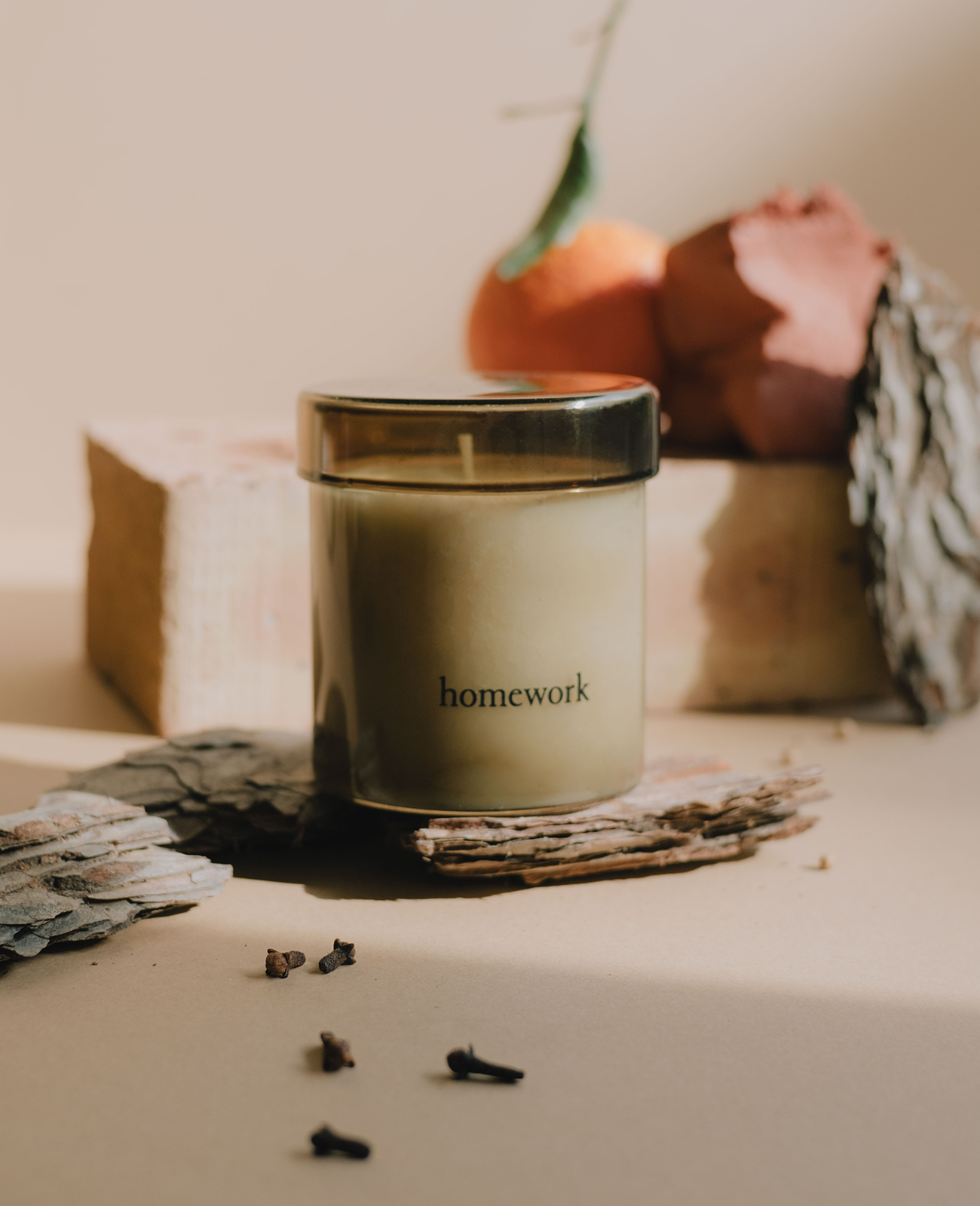 Homework fire candle in a moss green translucent glass with the branding homework written across the bottom in lower case lettering. On a grey background with cedar wood, kindling, star anise and oranges in the background.