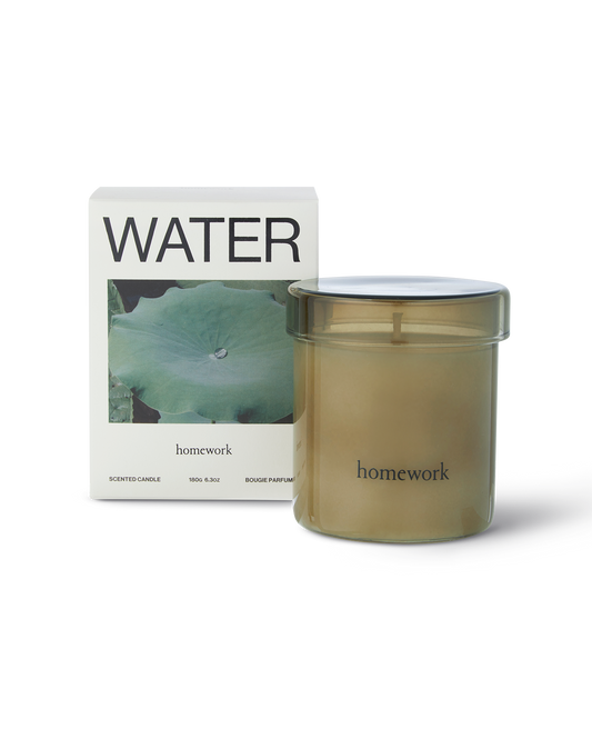 Homework water candle in a moss green cylindrical transparent glass with matching lid, along the bottom small branding spelling in lower-case lettering is homework. Next to the candle is the packaging a box with water written along the top, under is a landscape image of a green waterlily pad with a single water droplet on it. And under is the brands name homework in smaller lowercase lettering