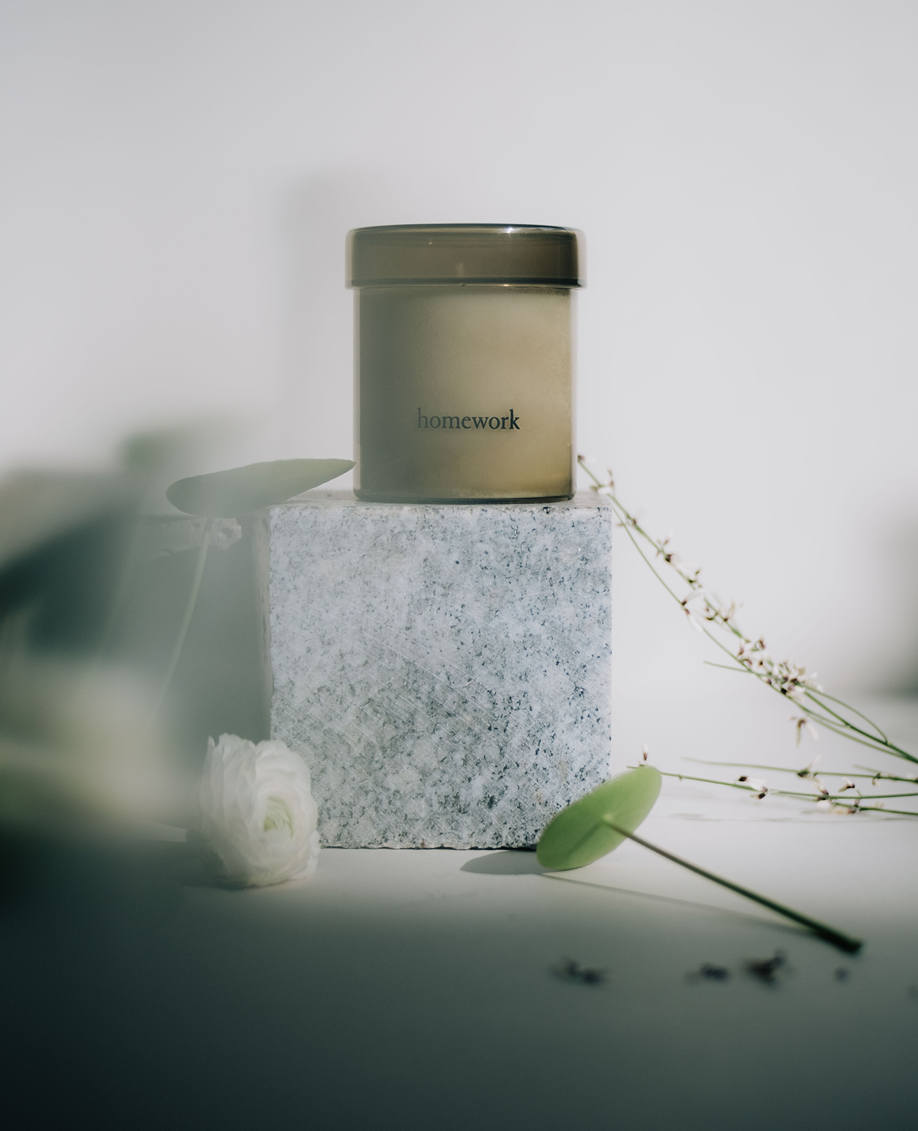 Homework Water candle in a moss green cylindrical translucent glass with matching lid. On a white backdrop, placed on top of a cube of marble, next to a white rose dud, two small lily pads and a few strands of jasmine plant