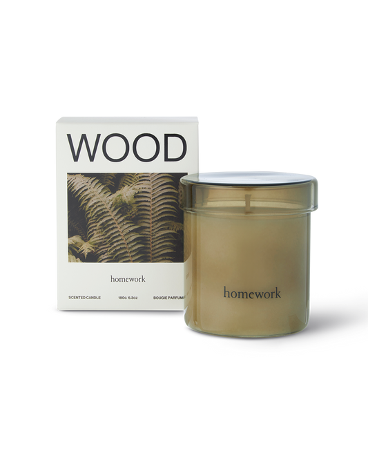 Homework wood scented candle in a moss green cylindrical glass with matching lid. Next to the packaging a white box with wood spelt across the top, under is a landscape image of a brownish green Fern plant. Under this has the branding lower case lettering of homework 