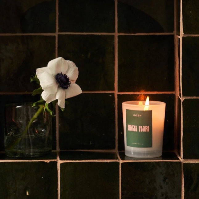 A candle with a green label with pink writing, sits against a black tiled wall next to a white flower