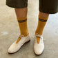 Person wearing mustard yellow socks with black stripes and white shoes on a concrete floor.