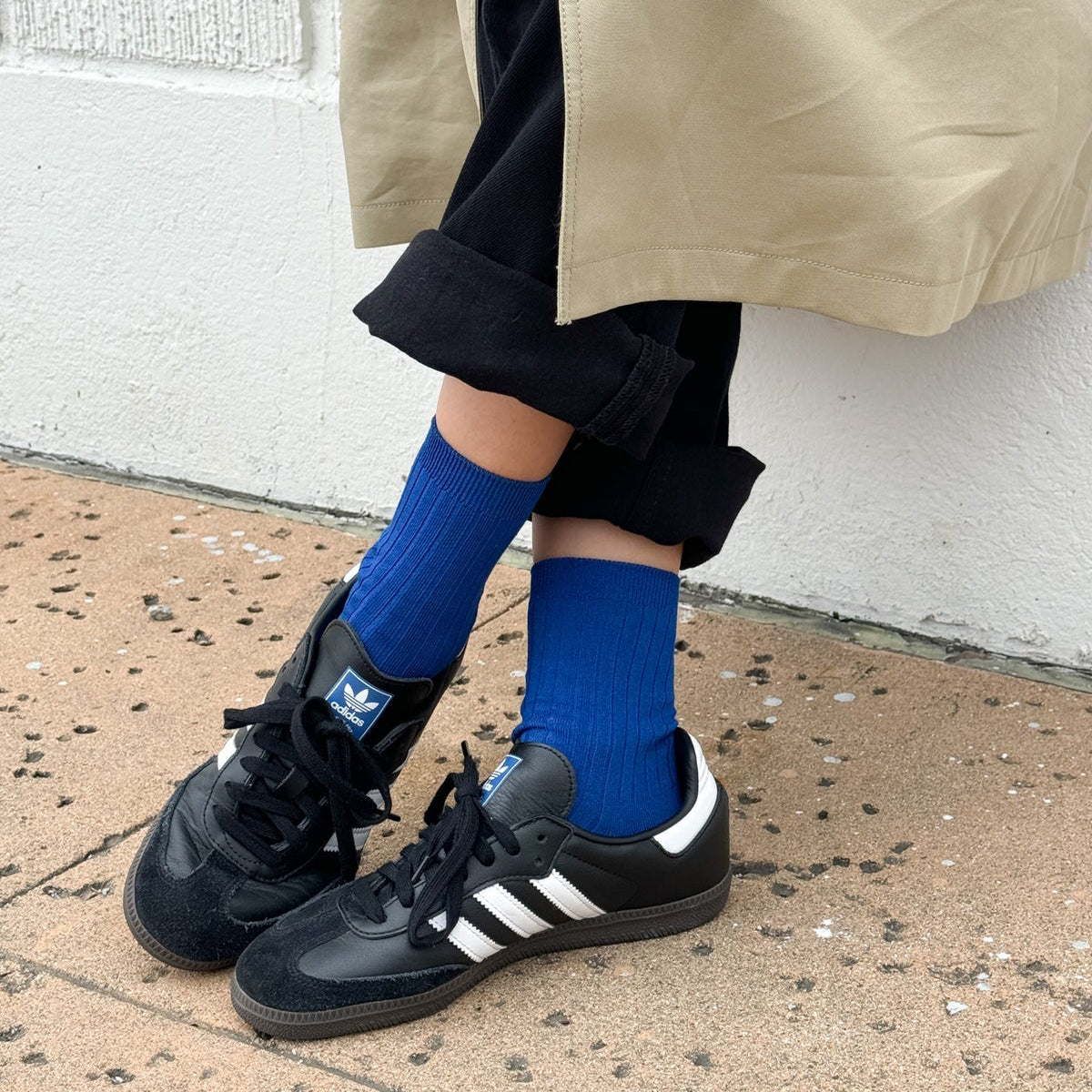 Person wearing black Adidas sneakers, blue socks, and a beige coat against a white wall.