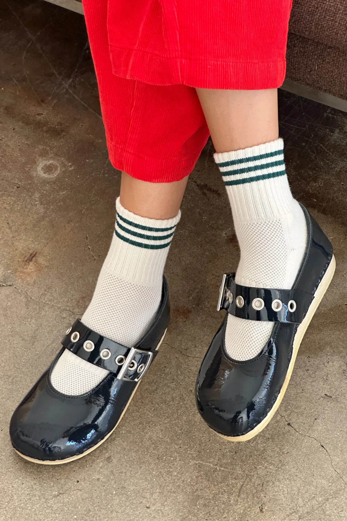 Person wearing navy blue shoes with buckles, white socks with green stripes, and red pants on a concrete floor.