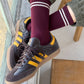 Person wearing maroon socks with white stripes and black sneakers with yellow stripes on a wooden floor.