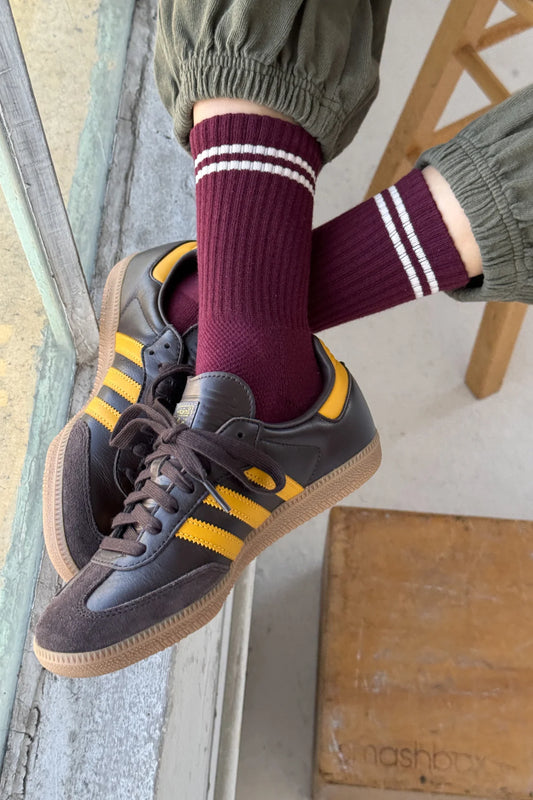 Person wearing maroon socks with white stripes and black sneakers with yellow stripes on a wooden floor.