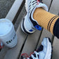 Person wearing white sneakers with black and blue accents on a wooden bench, next to a white cup.