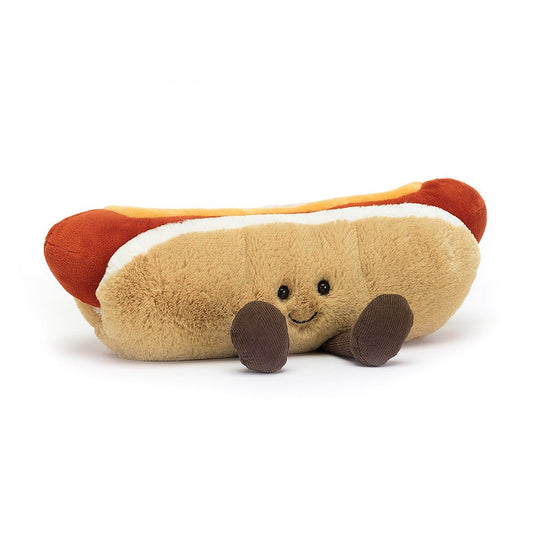 A plush toy in the shape of a hot dog. The hot dog is beige and has two brown legs coming out the bottom of the hot dog. The hot dog bun has a black smile and two black eyes, which appear to be made of beads. Inside the bun is a red sausage and on top of it is yellow mustard. This is against a white background.  