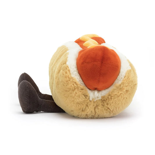 The side of a plush toy in the shape of a hot dog. The hot dog is beige and white at the top representing the inside of the bun and has two brown legs coming out the bottom of the hot dog. Inside the bun is a red sausage and on top of it is yellow mustard. This is against a white background. 