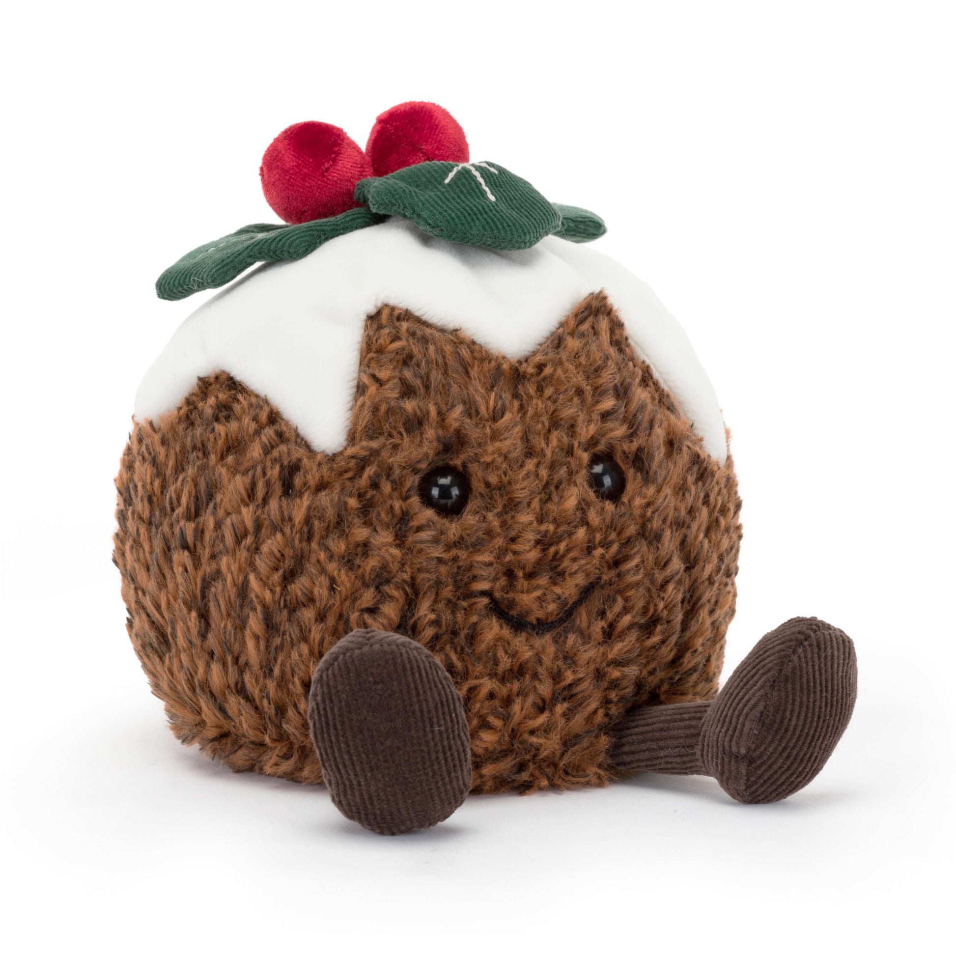 A plush Jellycat Amuseables Christmas Pudding with a textured brown body, white icing drizzle, two red berries, and a green leaf, sitting on a white surface. Shown in a jaunty side angle