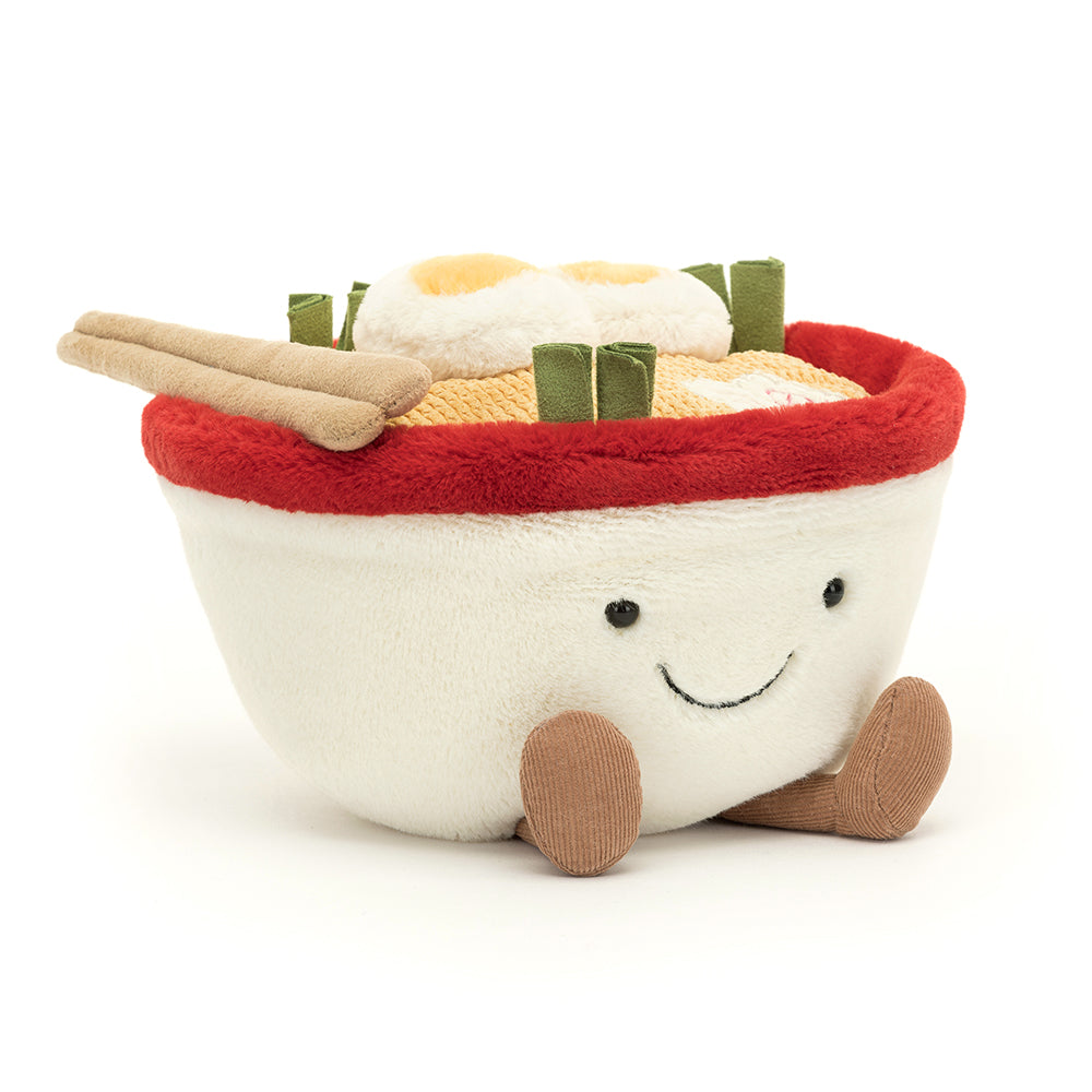 A quirky Jellycat Amuseables Ramen plush toy featuring a soft white bowl with a cheerful embroidered face, fluffy noodles spilling over the top, and vibrant plush toppings like an egg, seaweed, and a slice of pink swirl fish cake. Complete with corduroy legs for extra charm.