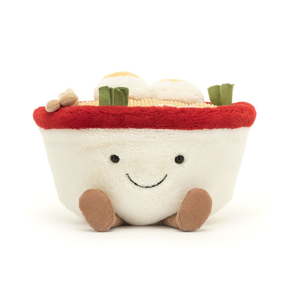 A quirky Jellycat Amuseables Ramen plush toy featuring a soft white bowl with a cheerful embroidered face, fluffy noodles spilling over the top, and vibrant plush toppings like an egg, seaweed, and a slice of pink swirl fish cake. Complete with corduroy legs for extra charm.