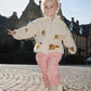 A young child wearing the Konges Sløjd Mon Grand Citron Jody Jacket walks along a cobbled street with arms outstretched. The cream-coloured fleece jacket features a playful lemon print, and the child pairs it with pink quilted trousers and patterned boots. A historic brick building is visible in the background under a bright sky.