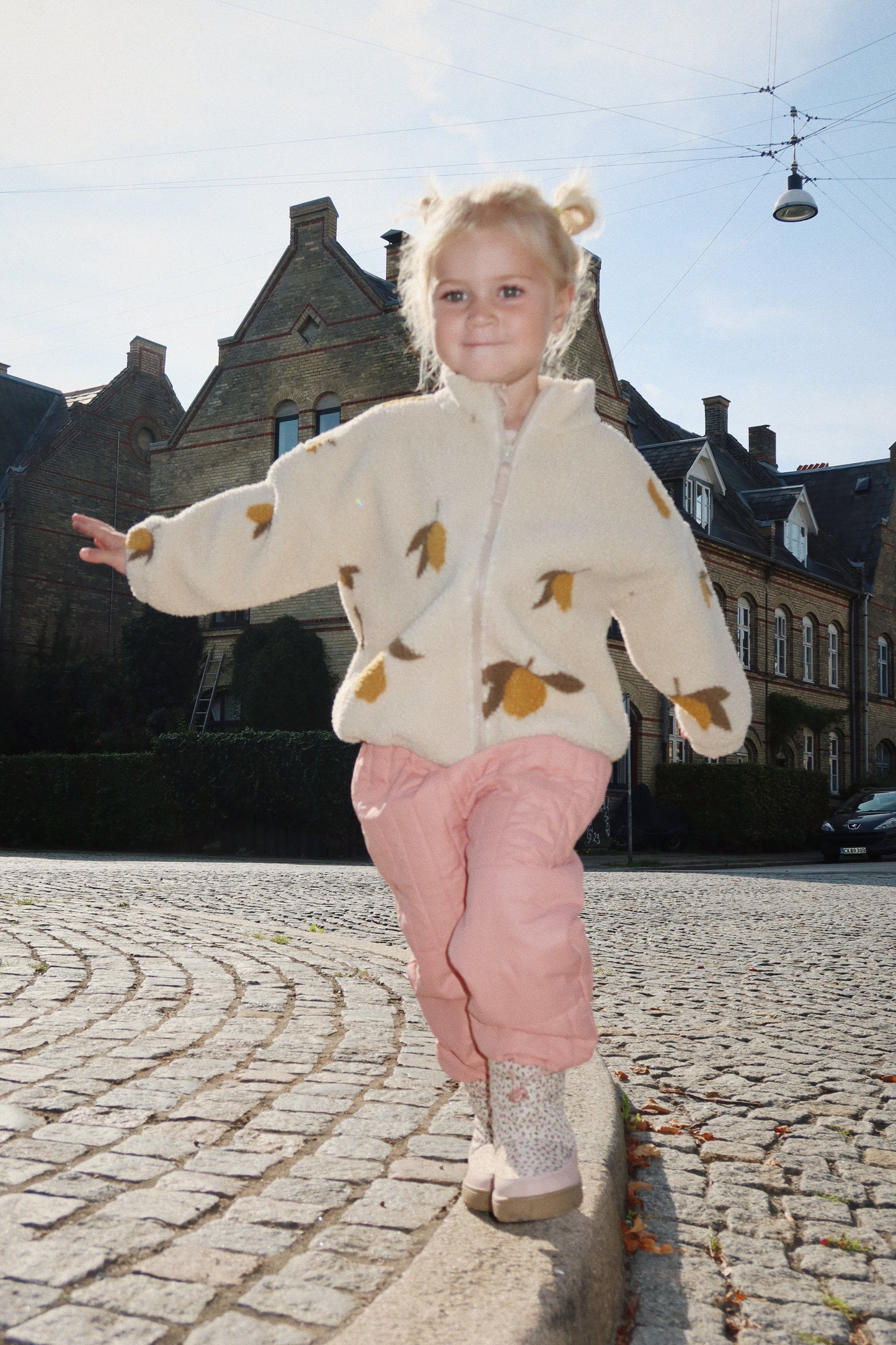 A young child wearing the Konges Sløjd Mon Grand Citron Jody Jacket walks along a cobbled street with arms outstretched. The cream-coloured fleece jacket features a playful lemon print, and the child pairs it with pink quilted trousers and patterned boots. A historic brick building is visible in the background under a bright sky.