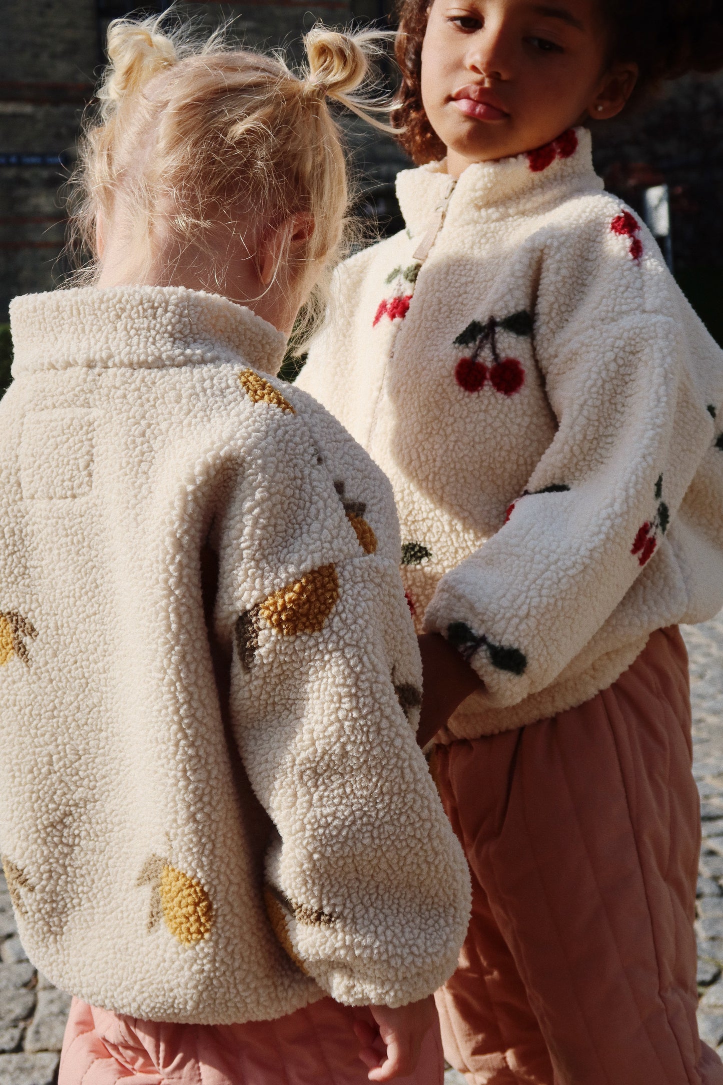 A child in the lemon-print Mon Grand Citron Jody Jacket and another in a cherry-print jacket stand close together outdoors.
