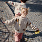 A child is walking on a cobbled street with arms outstretched, wearing the lemon-print fleece jacket, pink quilted trousers, and boots
