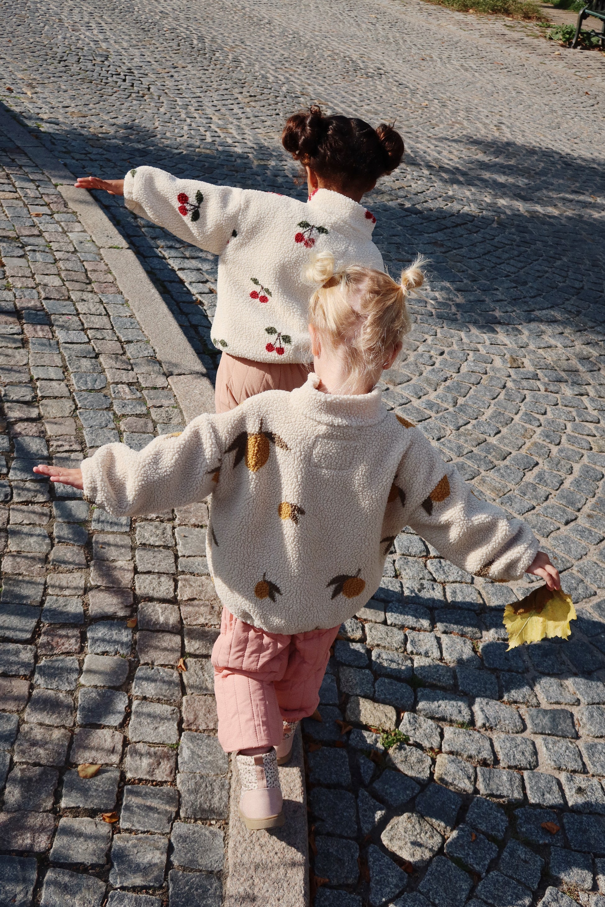 A child is walking on a cobbled street with arms outstretched, wearing the lemon-print fleece jacket, pink quilted trousers, and boots