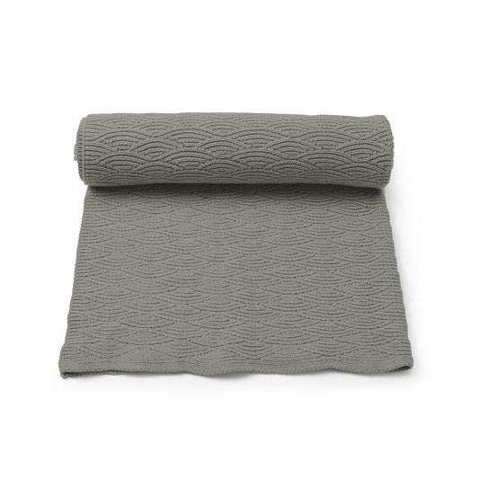 The Konges Sløjd Mulled Basil Pointelle Blanket, rolled up and partially unrolled, showcasing its delicate knit pattern in a muted green shade.