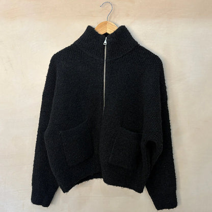 Black zip-up sweater on a wooden hanger against a beige wall.