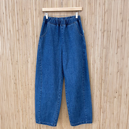 A pair of stylish high-waisted Arc Pants made from heavyweight denim, featuring a cropped length and slightly curved leg shape. The design includes unique side panels and deep pockets, perfect for casual wear. Displayed with a neutral background to highlight the pant's versatile style. They are hung on a wooden hanger against a wooden panel.