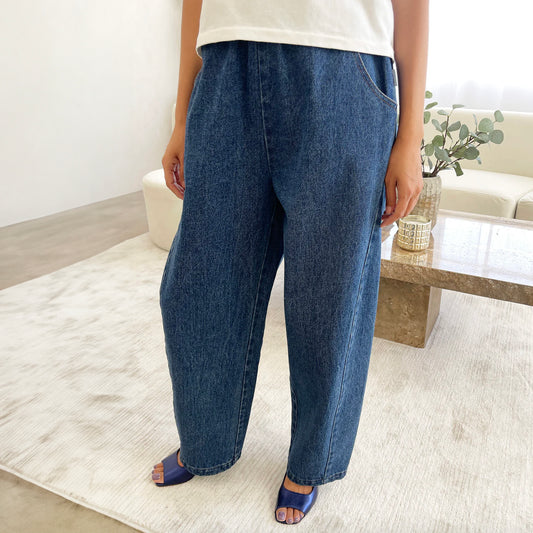 A pair of stylish high-waisted Arc Pants made from heavyweight denim, featuring a cropped length and slightly curved leg shape. The design includes unique side panels and deep pockets, perfect for casual wear.  They are worn with a white t-shirt and blue open toed sandals