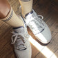 A pair of beige ribbed socks with navy blue stripe details at the cuff, styled with white sneakers on a wooden floor, illuminated by warm natural light.