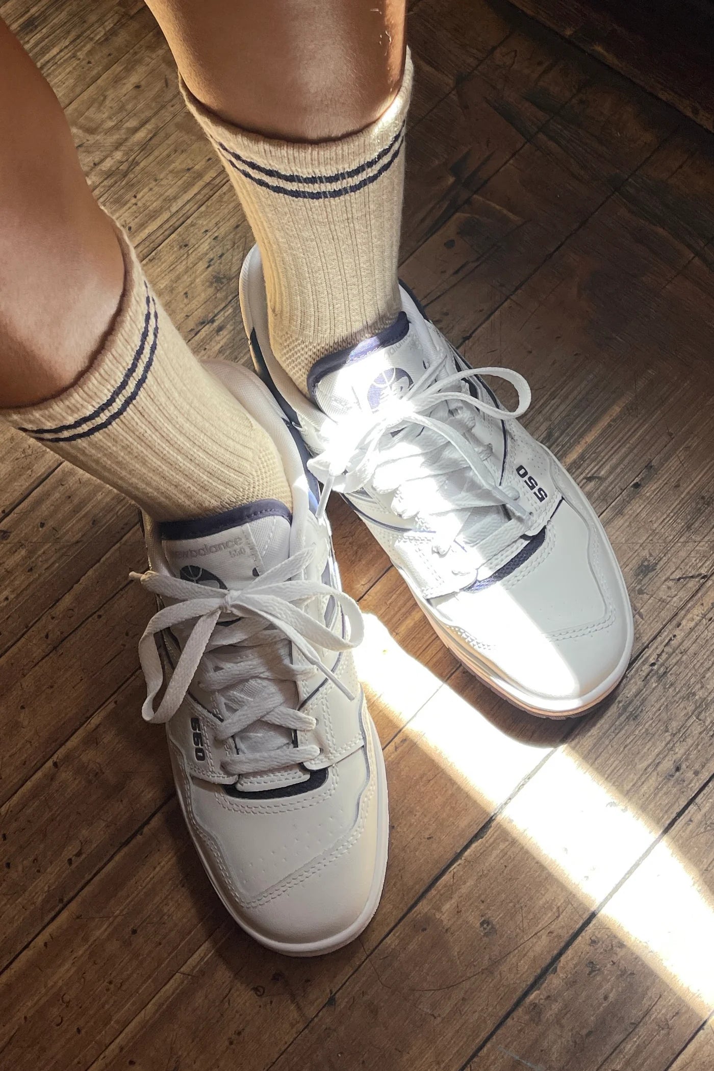 A pair of beige ribbed socks with navy blue stripe details at the cuff, styled with white sneakers on a wooden floor, illuminated by warm natural light.