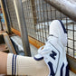 A close-up shot of a beige ribbed sock with navy blue stripe details at the cuff, worn with a white and navy sneaker, resting against a metal railing in an industrial setting.