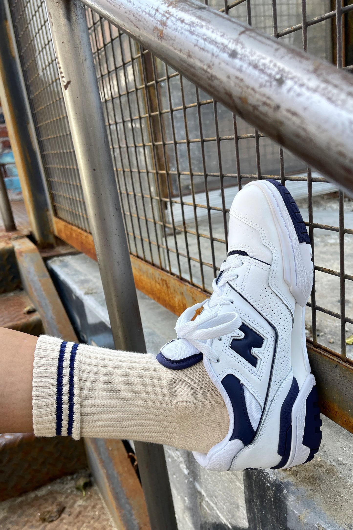 A close-up shot of a beige ribbed sock with navy blue stripe details at the cuff, worn with a white and navy sneaker, resting against a metal railing in an industrial setting.