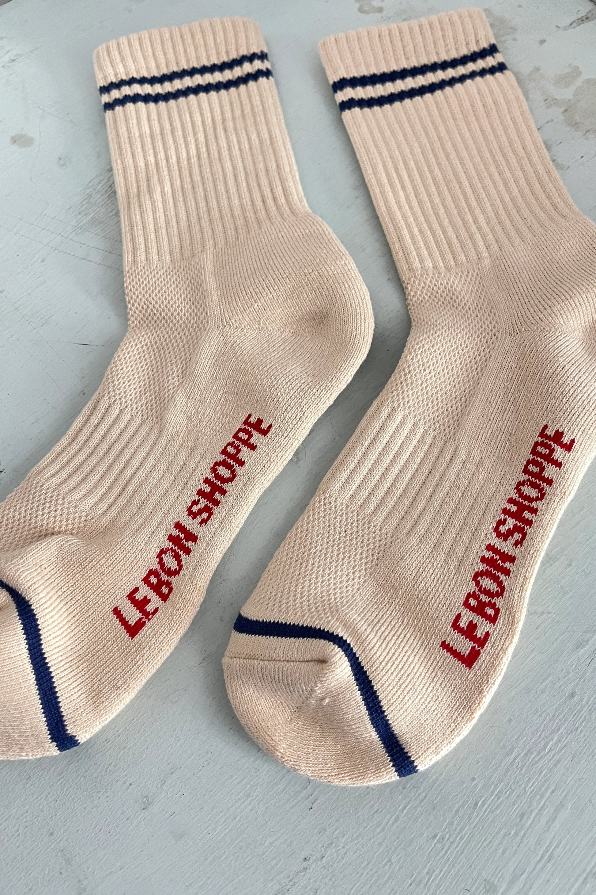 A pair of beige ribbed socks with navy blue stripe details at the cuff, displayed on a neutral surface, featuring red "Le Bon Shoppe" branding on the sole and navy blue contrast toe trim.