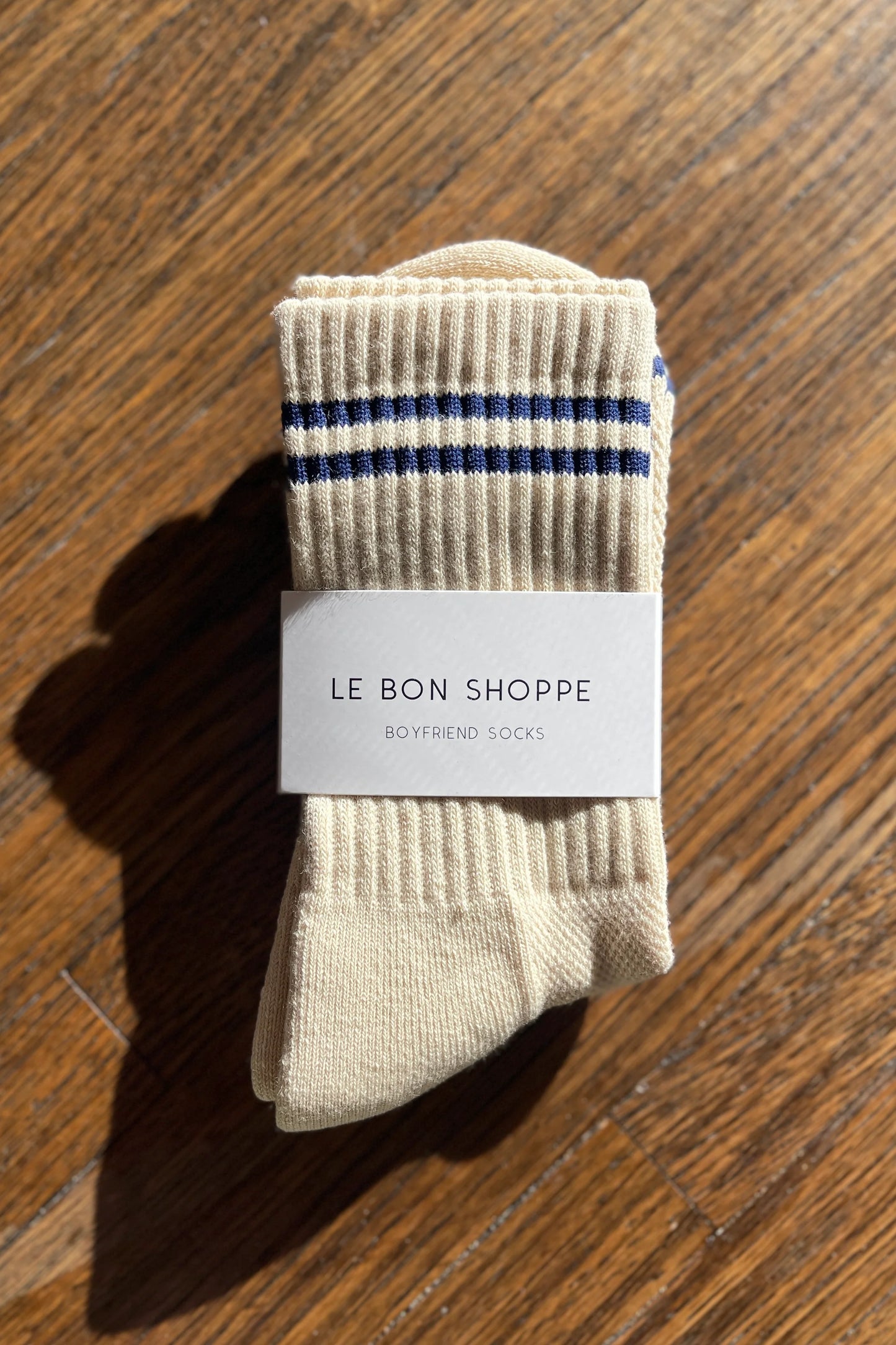 A neatly folded beige ribbed sock with navy blue stripe details at the cuff, wrapped in Le Bon Shoppe packaging, set on a wooden surface with natural light creating soft shadows.