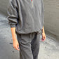 Person wearing a grey quarter-zip sweatshirt with a high collar, paired with matching sweatpants, standing on a concrete sidewalk in front of a textured grey wall.