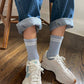 A person wearing light blue ribbed socks with white stripes at the cuffs, paired with rolled-up denim jeans and white sneakers, sitting on a wooden floor.
