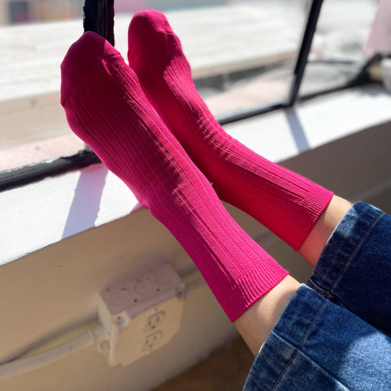 A pair of fuchsia ribbed socks worn with cuffed jeans, feet resting near a window with natural light streaming in.