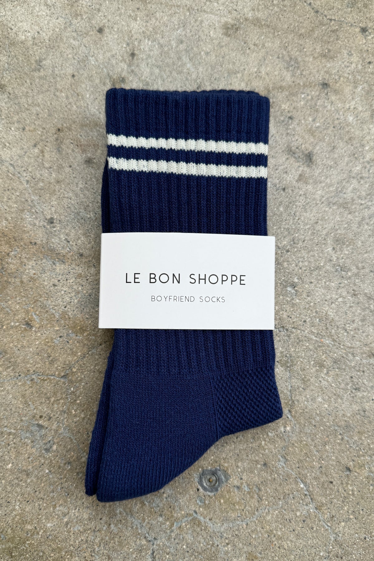 A pair of navy blue ribbed crew socks with two white stripes near the cuff, neatly folded and wrapped with a white Le Bon Shoppe label, placed on a textured concrete background.