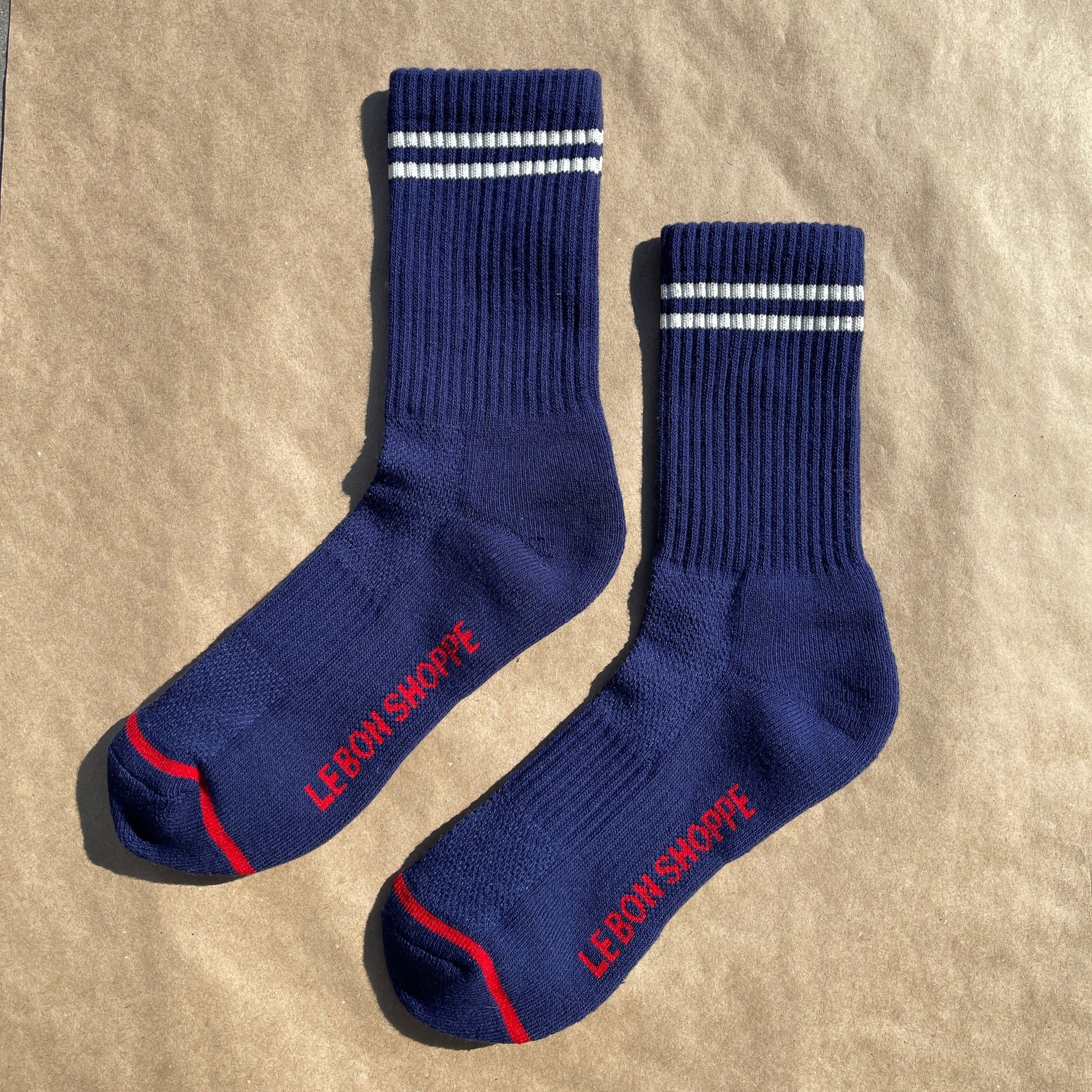 A pair of navy blue ribbed crew socks with white stripes near the cuff, laid flat on a brown paper background, showing red "Le Bon Shoppe" branding on the sole and red toe detailing.