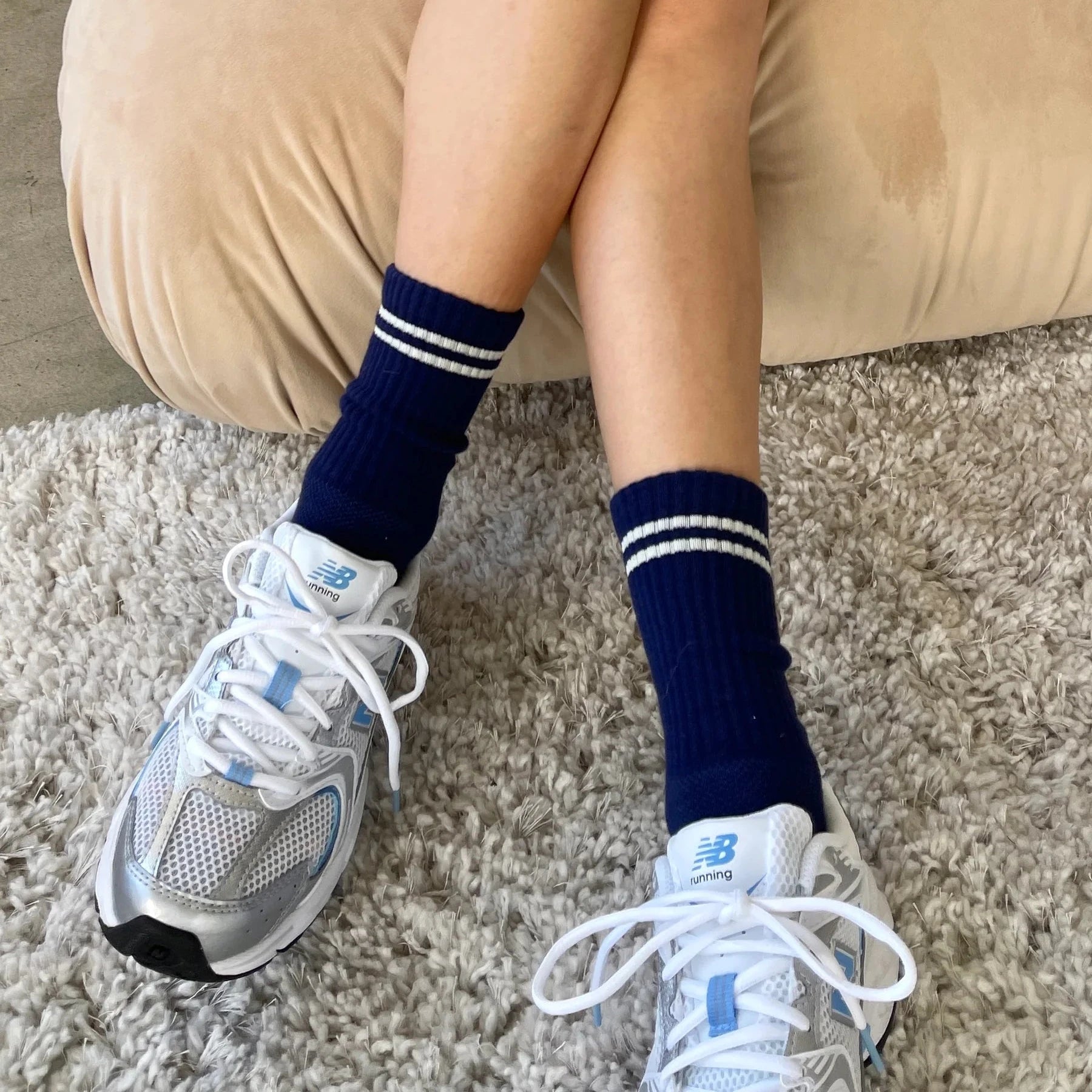 A person sitting on a beige cushion, wearing navy blue ribbed crew socks with white stripes, paired with white and blue New Balance sneakers, on a textured rug.