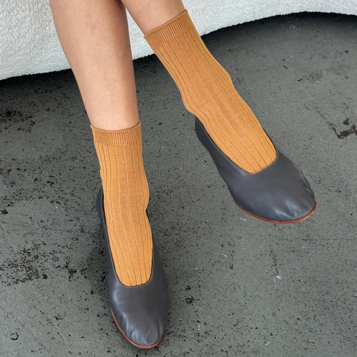 A person sitting on a textured bench, wearing peanut butter brown ribbed crew socks paired with black leather ballet flats, against a grey concrete floor.