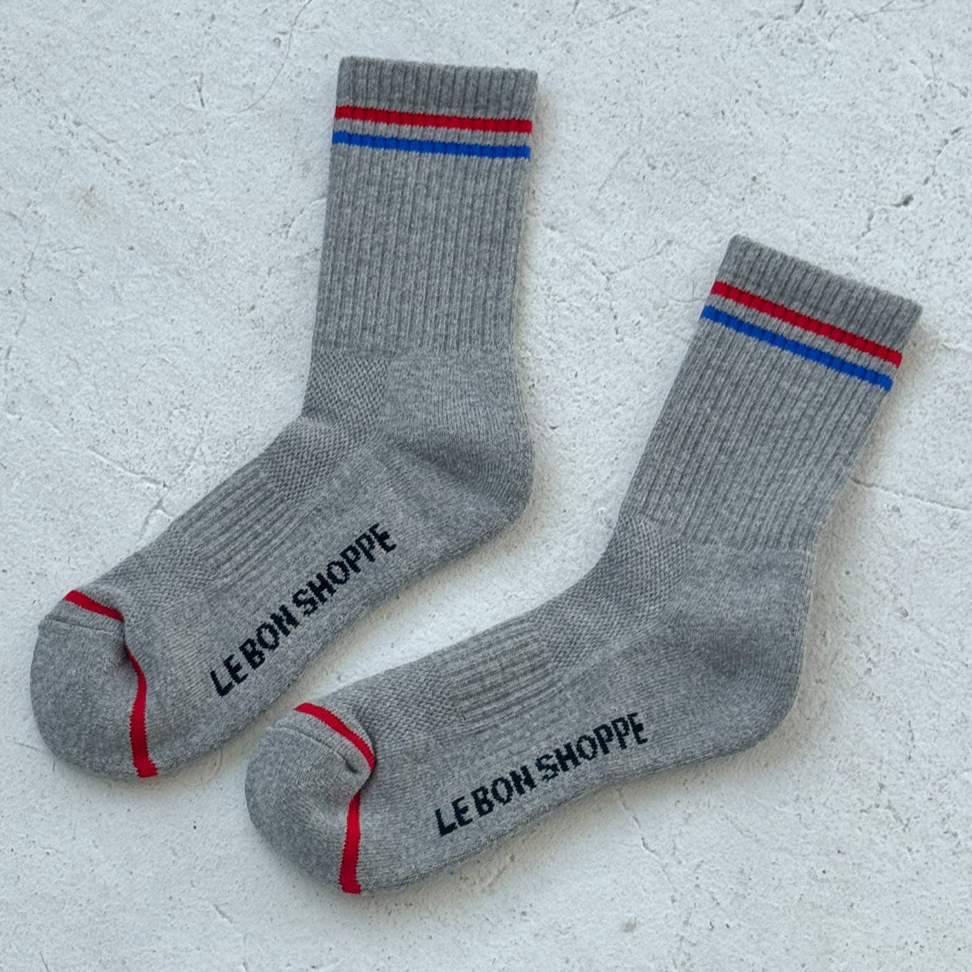 A pair of grey ribbed crew socks with red and blue stripes at the cuff, featuring "LE BON SHOPPE" branding on the sole, laid flat on a textured light grey surface.