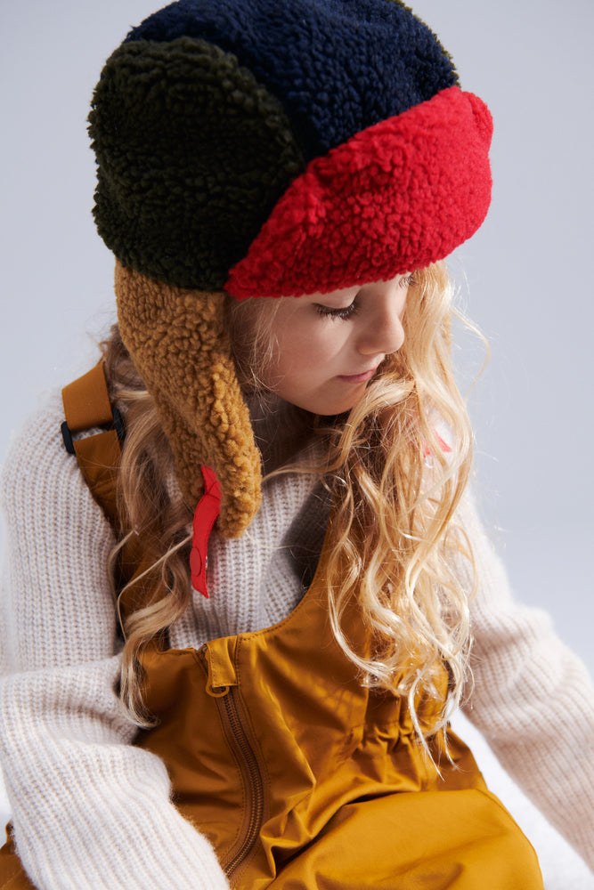 The model with long blonde wavy hears wears the Liewood Bravo Pile Hat in Army Brown/Multi Mix, featuring brown, navy blue, and red color tones, with soft fleece lining, earflaps, and adjustable snap-button straps. She wears a cream jumper and tan dungarees