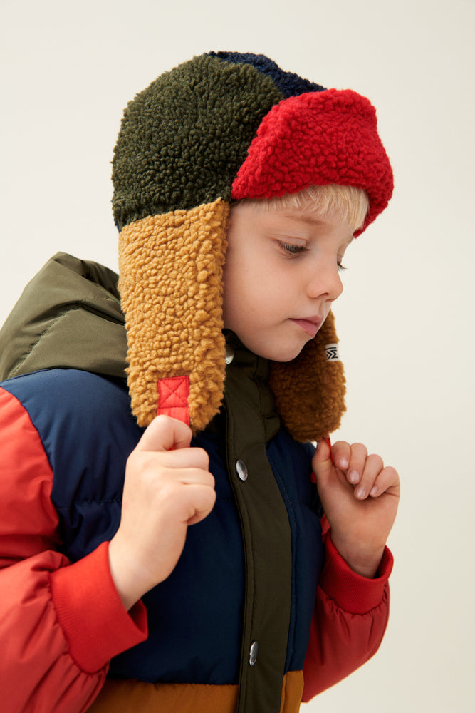 The model with short blonde hair wears the Liewood Bravo Pile Hat in Army Brown/Multi Mix, featuring brown, navy blue, and red color tones, with soft fleece lining, earflaps, and adjustable snap-button straps. He wears a matching padded jacket