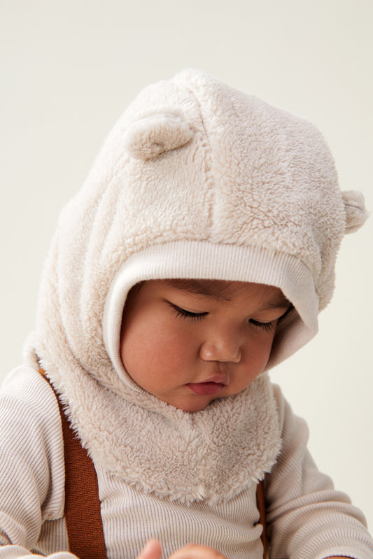 Liewood Giri Balaclava in soft teddy fabric, featuring ear details, a ribbed hem around the head opening, and a small bunny embroidery, worn on a young toddler wearing a cream rib top and cinnamon braces