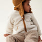 Liewood Golden Caramel Bravo Pile Hat for children, featuring soft fleece lining, protective earflaps, and adjustable snap-button straps. The model looks to the side in a cream  fleece jacket and beige cords