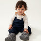 A toddler with dark curly hair and a cute grumpy face wears Liewood Heather Baby Footies in Mini Leo/Mist, featuring a soft fleece lining, water-repellent and windproof material, Velcro ankle closure, and non-slip soles, displayed on a neutral background.