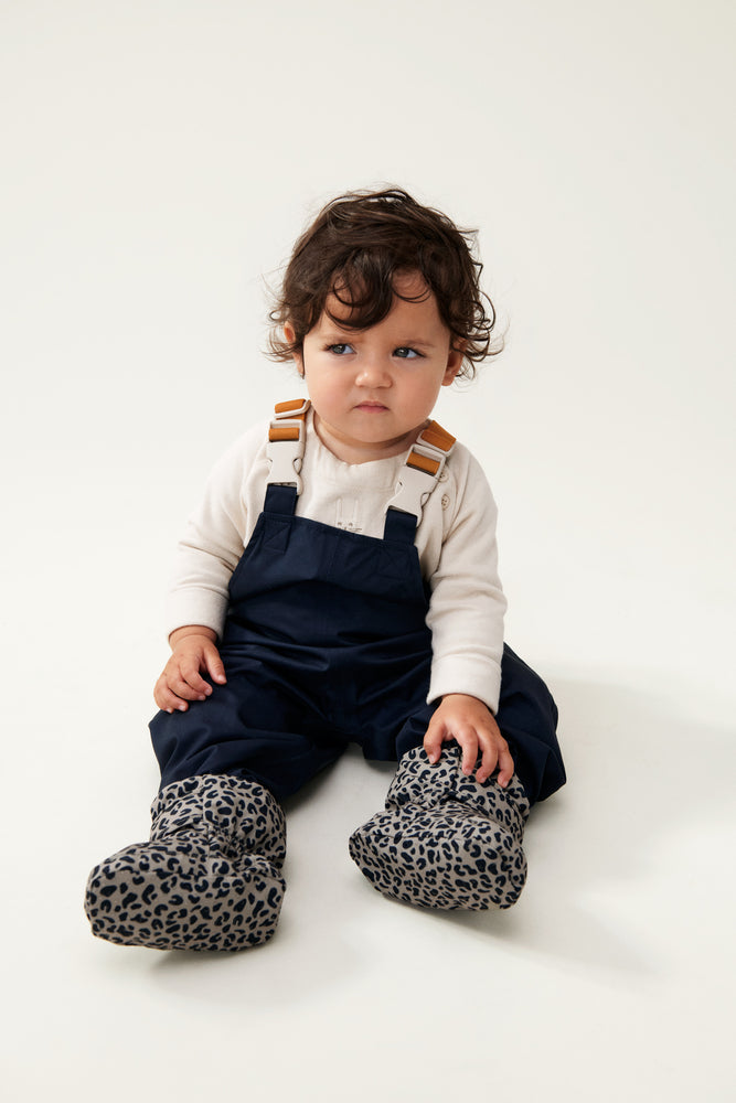 A toddler with dark curly hair and a cute grumpy face wears Liewood Heather Baby Footies in Mini Leo/Mist, featuring a soft fleece lining, water-repellent and windproof material, Velcro ankle closure, and non-slip soles, displayed on a neutral background.