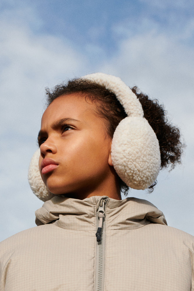 The model wears Liewood Albero Pile Ear Warmers in Sandy, featuring soft recycled fleece, a neutral sandy colour, and a cozy fit. She is wearing a beige zip up jacket and stands against a blue sky